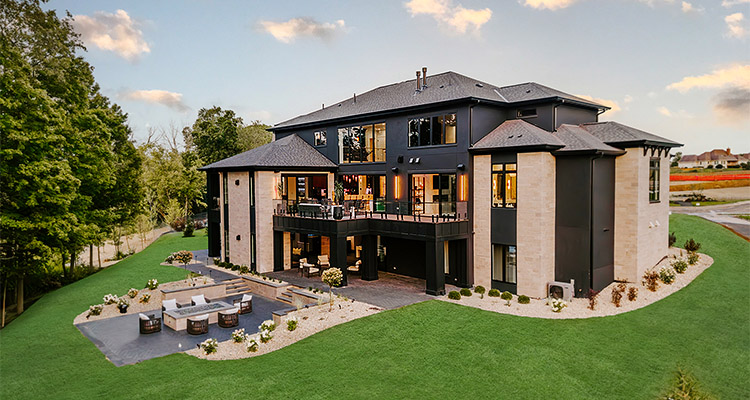 large modern detached residential home featuring a mix of dark exterior panels and light-colored stone, situated on a well-manicured lawn with a patio and fire pit area