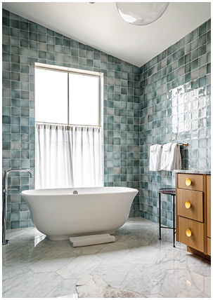 a modern bathroom design showcasing teal or blue-green crackle-effect wall tiles paired with a white freestanding bathtub and light-colored marble flooring. 