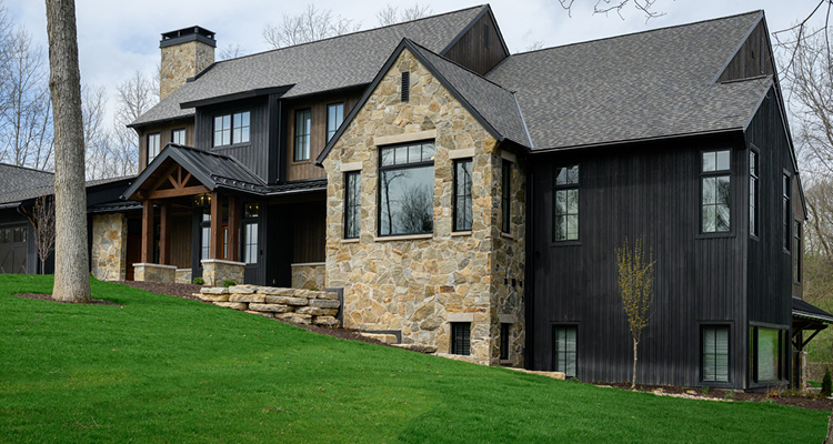 a large, modern farmhouse with a striking mixed-material exterior. The home sits on a sloped green lawn and utilizes a high-contrast color palette.