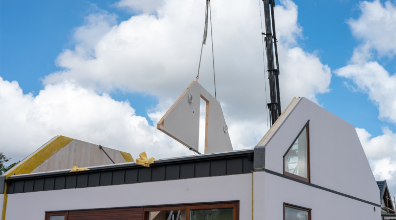 Crane lifting prefabricated wall panel onto modern modular house under construction.
