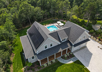 An aerial, high-angle view shows a large, two-story luxury home surrounded by thick green woods