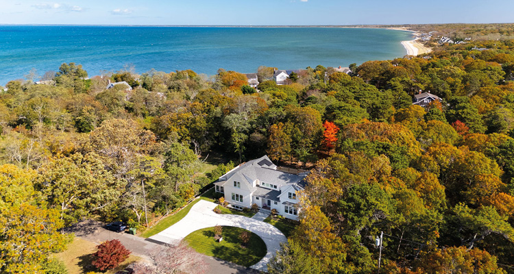 An aerial, high-angle photograph captures a large, modern two-story white house nestled within a dense forest of autumn-colored trees
