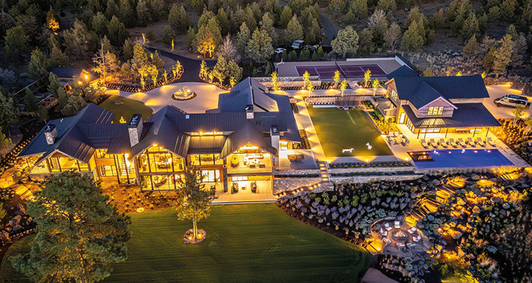 An aerial drone photograph captures a sprawling luxury estate at dusk, brilliantly illuminated by warm exterior accent lighting