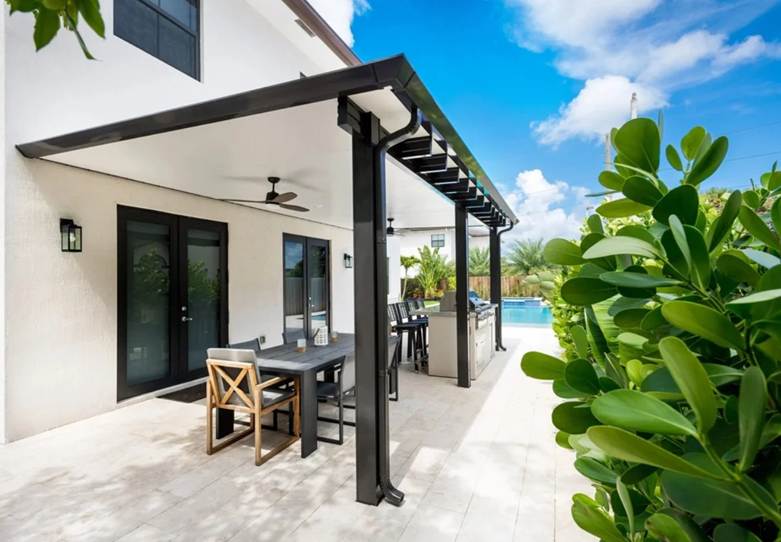 Black aluminum pergola with open louver design extending from a residential exterior. The structure features clean lines and connects the home to an outdoor living area near a pool.