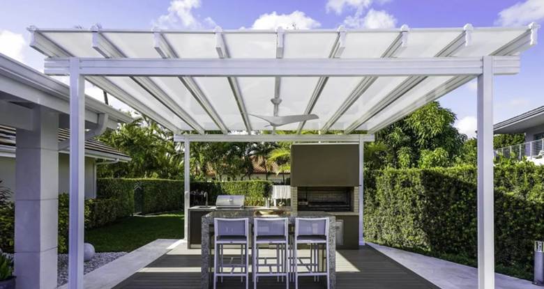 White louvered pergola sheltering an outdoor kitchen area with adjustable roof slats. The aluminum structure features a ceiling fan and provides coverage for built-in cooking and dining spaces.