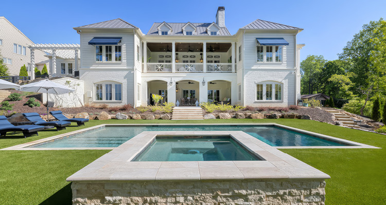 the rear exterior of a luxury white two-story traditional-style residential home with a resort-style swimming pool and raised stone spa