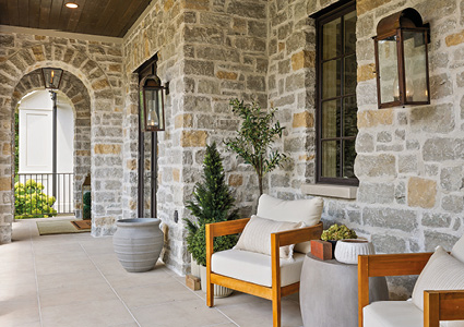 a covered outdoor patio with a rustic stone wall, wooden-framed white cushion armchairs, and black metal wall lanterns