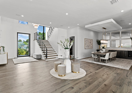 a bright, open-plan modern living and dining area featuring light wood flooring, a central round accent table with four upholstered stools, a staircase with a black metal railing, and a formal dining set with white chairs