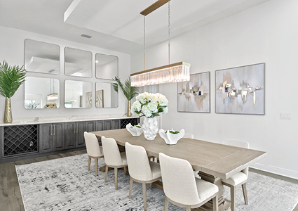 a modern, bright dining room featuring a long wooden table, white upholstered chairs, a linear crystal chandelier, a mirrored wall accent, and a gray sideboard with a wine rack