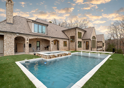 two-story brick residence with a gabled roof, an attached covered patio, a raised stone hot tub, and a rectangular in-ground swimming pool