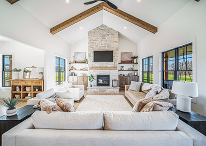 a modern farmhouse-style living room featuring a vaulted ceiling with exposed wood beams, a light stone or brick fireplace as the focal point, neutral-toned sectional seating, and large windows