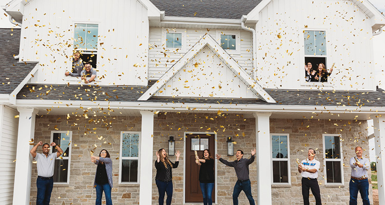 a group of people celebrating with gold confetti outside the front of a modern two-story house with stone and white-siding accents