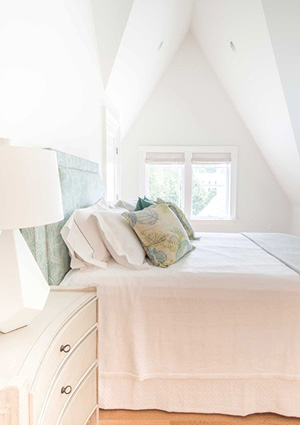 a bright, white attic bedroom featuring a light-toned upholstered headboard, white bedding with a bed skirt, a white nightstand with dark hardware, a tall white table lamp, and a small multi-colored accent pillow