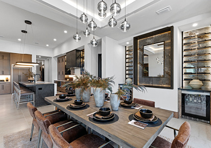 a modern open-plan dining and kitchen area featuring a large wooden dining table with upholstered armchairs, a central island with bar stools, cluster pendant lighting, and a built-in glass wine rack feature wall