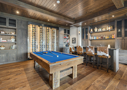a luxury rustic-modern home game room and bar featuring a blue-felt pool table with a light wood frame, a glass-enclosed wine wall, and a full bar with cowhide-upholstered barstools