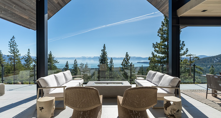 a luxurious, modern outdoor patio overlooking a panoramic view of Lake Tahoe surrounded by pine trees and snow-capped mountains under a clear blue sky
