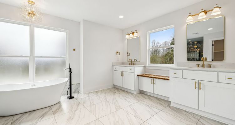 a modern, bright bathroom with white double vanities, gold hardware, marble-look polished floor tiles, a freestanding soaking tub, and a black floor-mounted tub filler