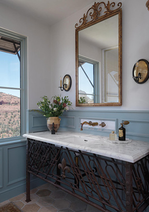 a rustic-industrial bathroom vanity with an ornate wrought-iron base, a white marble top, and a matching antique-style scroll frame mirror
