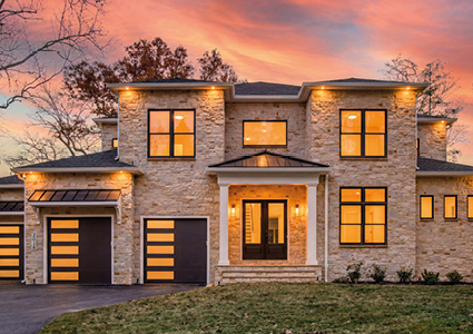 a modern, two-story stone-clad luxury single-family house with dark-framed windows, integrated garage doors, and a dramatic sunset sky.